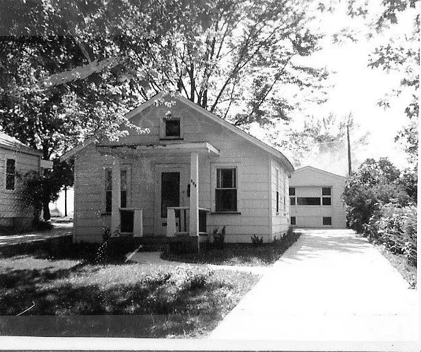 Black-and-white photo of a small single-story house with a front porch, a driveway leading to a detached garage, and large trees behind the house.