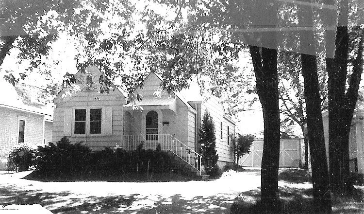 Black-and-white photo of a small single-story house with a front porch and stairs, shaded by large trees, with a driveway leading to a detached garage.