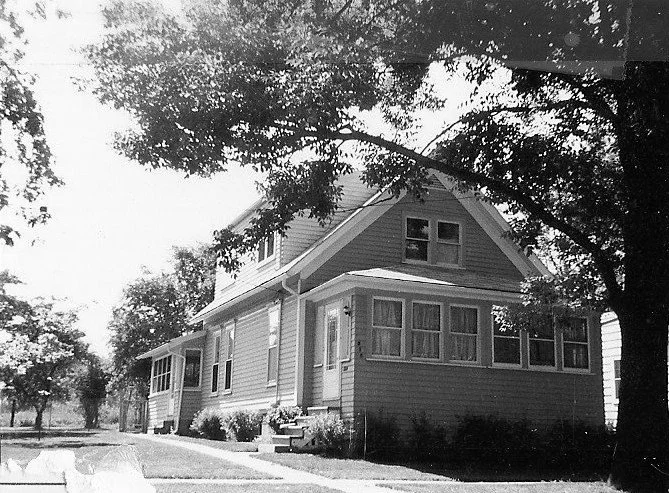 Black-and-white photo of a two-story bungalow with an enclosed front porch and a large tree in the foreground.