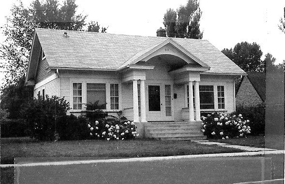Black-and-white photo of a single-story bungalow with a covered front porch supported by two columns, steps up to the door, and flowering shrubs along the front lawn.