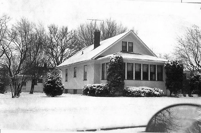Snow-covered bungalow with enclosed front porch, bare trees and shrubs, and a car mirror in the foreground.