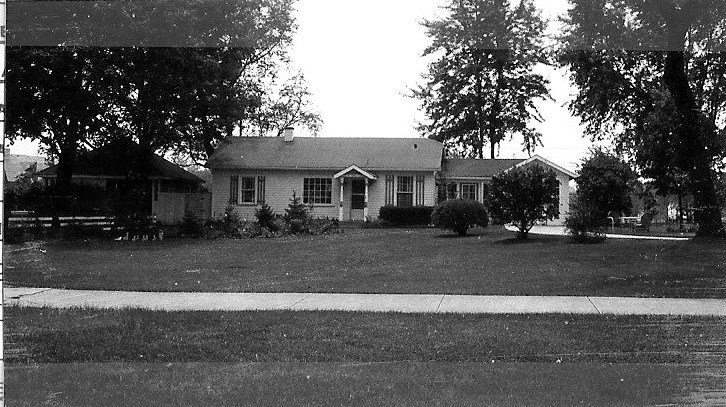 Black-and-white photo of a single-story ranch house with a wide front lawn, sidewalk, and large trees framing the property.