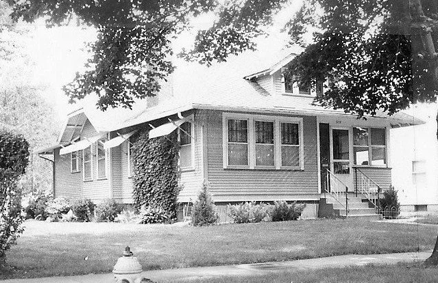 Black-and-white photo of a single-story bungalow with a front porch, window awnings, climbing ivy, lawn and a fire hydrant in the foreground.