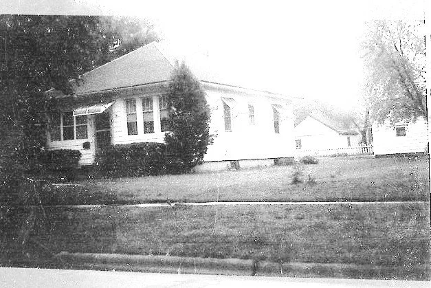Black-and-white photo of a single-story house with a small front porch, lawn, and trees.