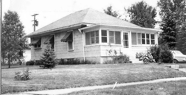 Black-and-white photo of a single-story bungalow with an enclosed front porch, lawn and shrubs, trees in the background and a car partially visible at right.