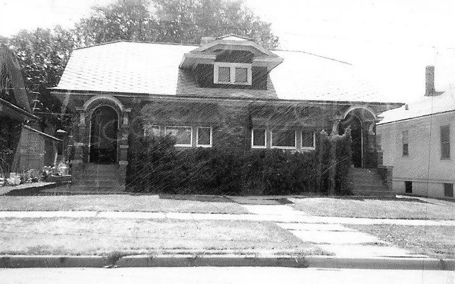 Front view of a one-story brick bungalow with a central dormer, two arched entryways, and a small front lawn.