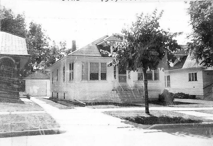 Black-and-white photo of a single-story bungalow with an enclosed front porch, small tree in the front yard, and a driveway leading to a detached garage.