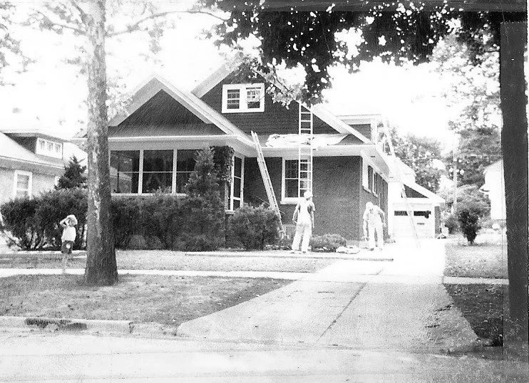 Black-and-white photo of a bungalow with two workers on ladders repairing the roof while a child stands on the sidewalk.