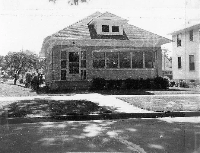 Black-and-white photo of a single-story brick house with an enclosed front porch, hipped roof dormer, and a sidewalk and lawn in front.