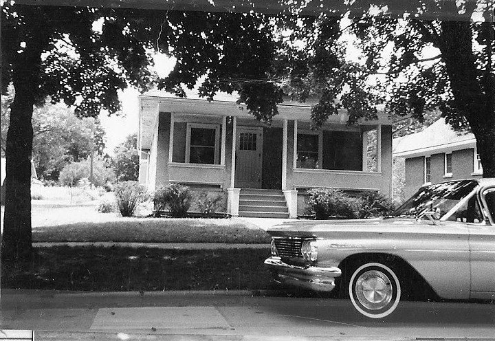 Black-and-white photo of a single-story bungalow with a front porch framed by large trees and the front end of a vintage car parked on the street.