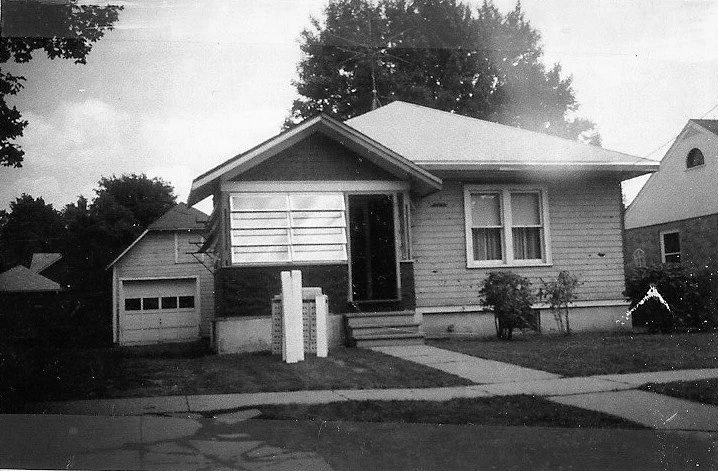 Black-and-white photograph of a small single-story bungalow with a detached garage, front steps and sidewalk, two mailbox posts on the lawn, and large trees behind the house.