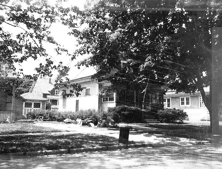 Black-and-white photo of a two-story house partly shaded by a large tree, with shrubs, front lawn, sidewalk and a curbside trash can.