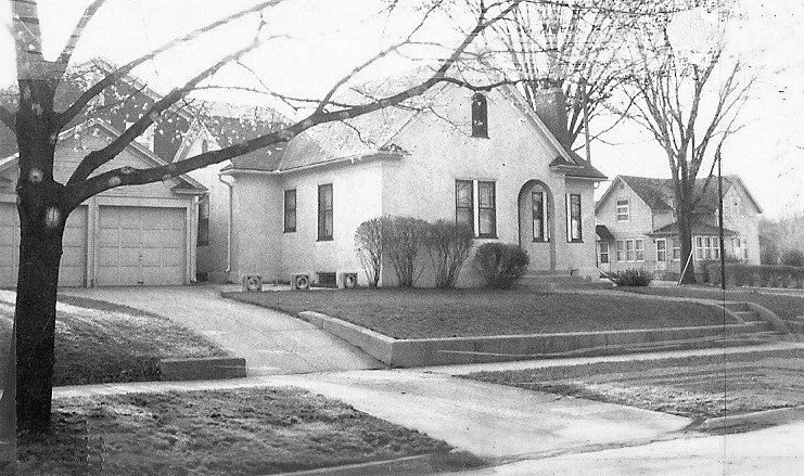 Black-and-white photo of a single-story stucco house with shrubs, a driveway to a two-car garage, leafless trees, and neighboring homes in a quiet residential street.
