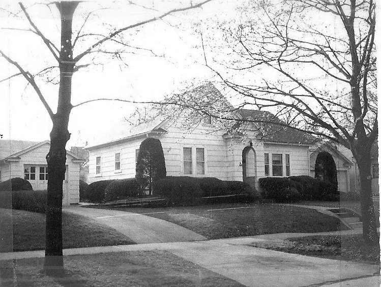 Black-and-white photo of a single-story suburban house with trimmed shrubs, a curved driveway, and leafless trees in the foreground.