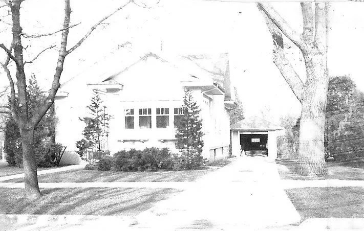 Black-and-white photo of a bungalow-style house with a front yard, driveway leading to a detached garage, and large trees framing the scene.