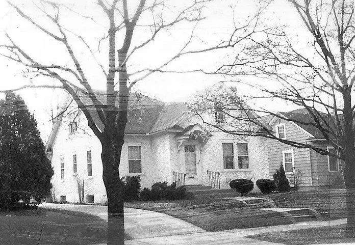 A black and white photo of a white stucco house with a prominent front porch, surrounded by bare trees and a sidewalk.