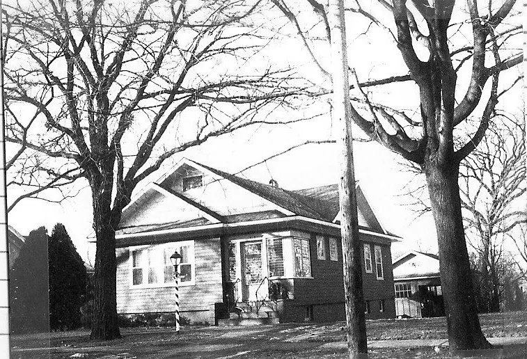 A black and white photo of a house framed by two large, bare trees, with a striped street lamp and a utility pole in the foreground.