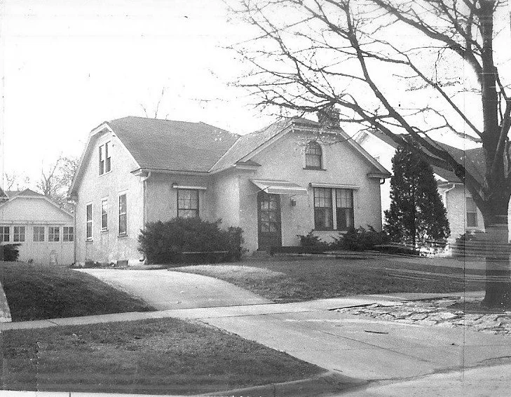 A black and white photo shows a stucco house with a driveway, a detached garage, and bare trees in the foreground and background.