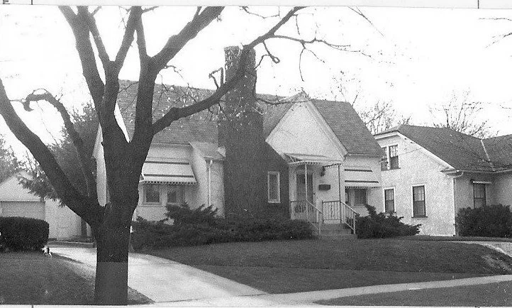 Black-and-white photo of a single-story bungalow with a central chimney and small front porch, a large leafless tree in the foreground, driveway and lawns, and neighboring houses visible.
