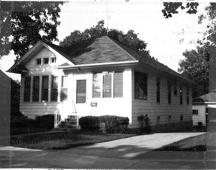 Black-and-white photo of a single-story wood-sided bungalow with an enclosed front porch, small front steps and shrubs, and a driveway along the right side.