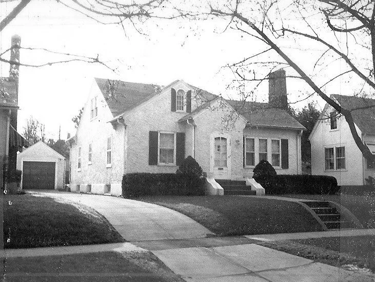 Black-and-white photo of a single-story stucco house with a front porch, detached garage at left, curved driveway, trimmed hedges, and leafless tree branches framing the scene.
