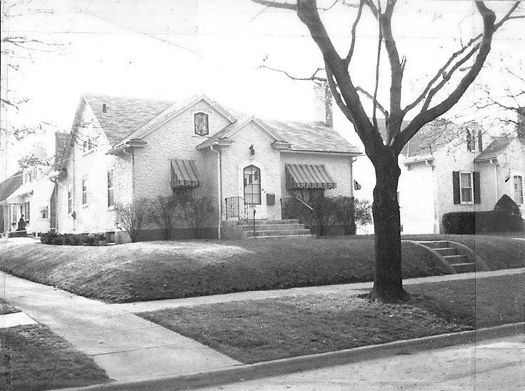 Black-and-white photo of a single-story cottage-style house with striped awnings, front steps and railing, a large tree in the foreground, and a sidewalk-lined residential street.