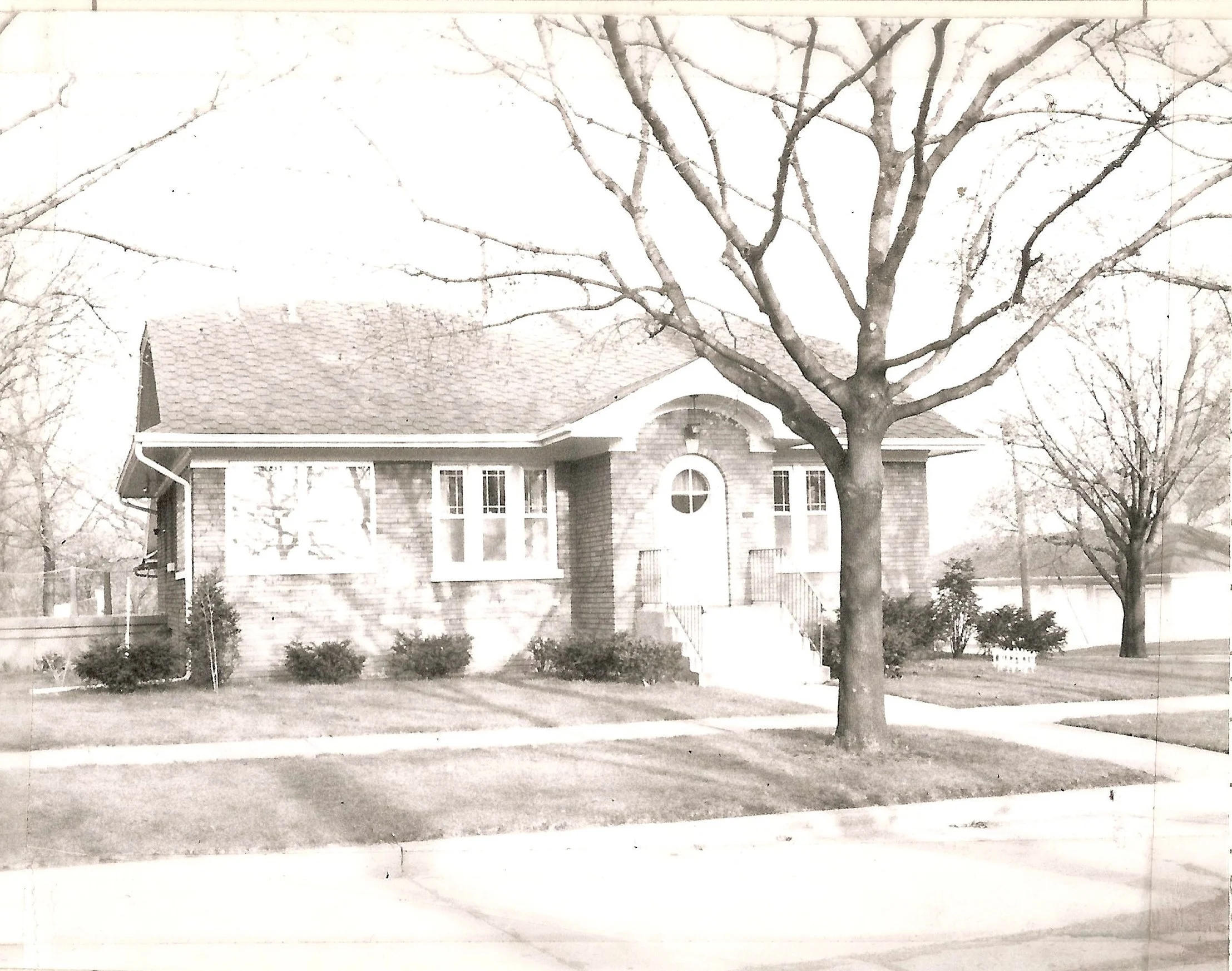 Black-and-white photo of a brick bungalow with a large leafless tree in front and a tidy lawn.