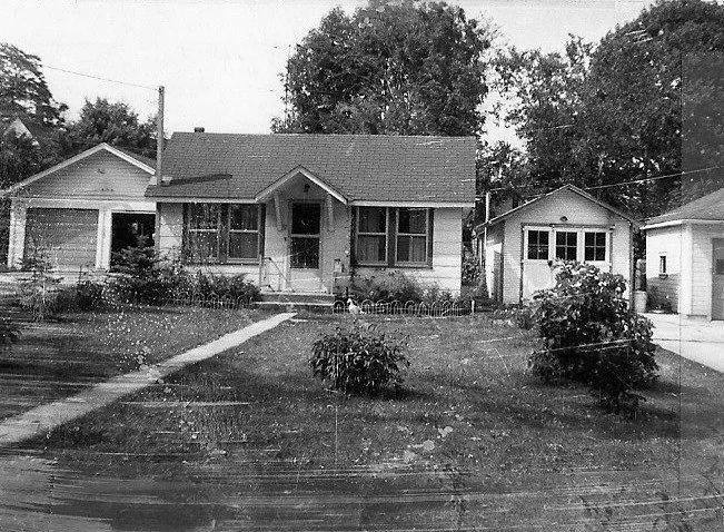 Black-and-white vintage photo of a small single-story house with a front porch, a straight walkway across the lawn, two detached garages at either side, and trees in the background.
