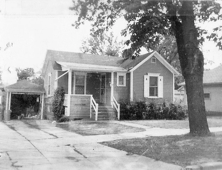 Small single-story bungalow with a front porch, steps, attached carport and large tree in the front yard.