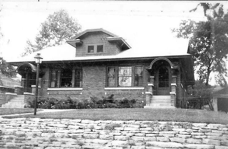Black-and-white photo of a single-story brick bungalow with a central dormer, arched front entry and steps, a lamp post, and a stone retaining wall.