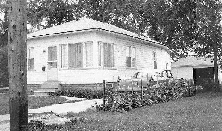 Single-story white bungalow with an enclosed front porch, two vintage cars parked beside it and a detached garage in the yard.