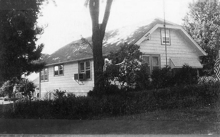 Black-and-white photo of a wood-sided house with a steep roof and dormer, trees and shrubs in front and an American flag visible at the right edge.