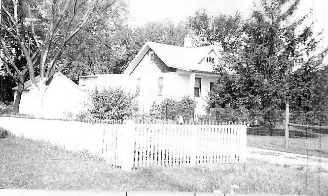 Black-and-white photo of a two-story house with a pitched roof, trees around it, and a white picket fence in the foreground.