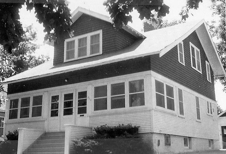 Black-and-white photo of a two-story house with an enclosed front porch, dormer windows, and tree branches framing the roof.