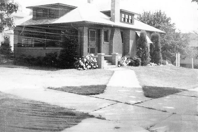 Black-and-white photo of a bungalow-style house with a front walkway, steps, shrubs, and a small porch.