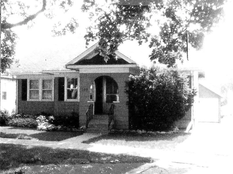 Black-and-white photo of a small brick bungalow with arched front porch, steps, large tree and driveway leading to a detached garage.