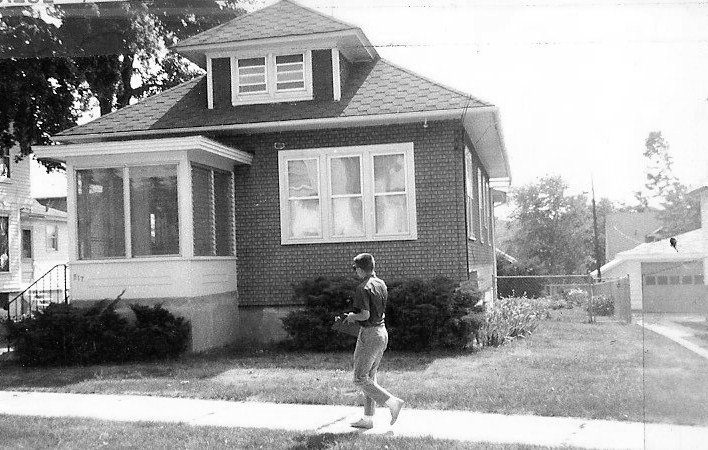 Black-and-white photo of a person walking on a sidewalk past a brick bungalow with an enclosed front porch and a detached garage in the background.