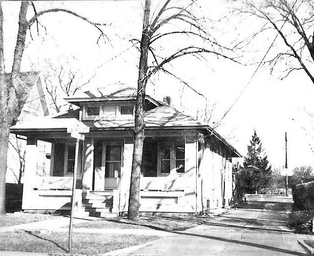 Black-and-white photo of a one-story bungalow with a covered front porch and steps, two leafless trees in front, a street sign at the corner and a driveway along the right side.