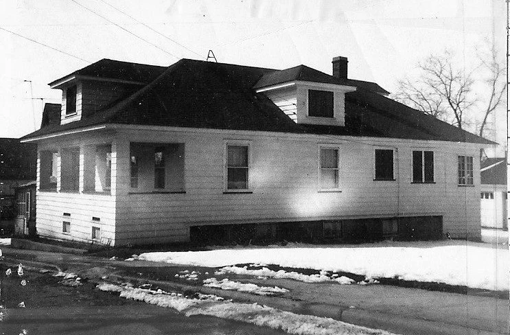 Black-and-white photo, side view of a single-story bungalow with dormer windows, clapboard siding, and patches of snow on the yard and driveway.