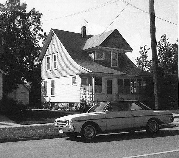 Black-and-white photo of a two-story wood-frame house with a covered porch and a vintage two-door car parked on the street in front.