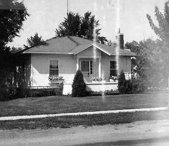 Black-and-white photograph of a single-story bungalow with a small front porch, window flower boxes, a manicured lawn, and trees in the background.