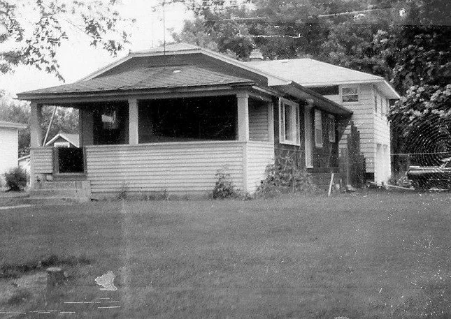 Black-and-white photo of a single-story house with a covered front porch, steps at left, and a grassy lawn in front.