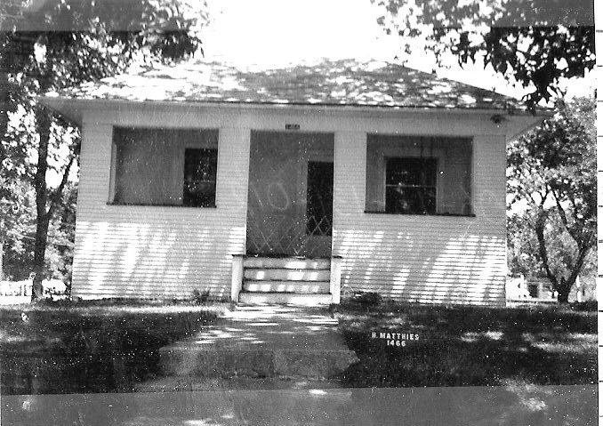 Black-and-white photo of a small single-story bungalow with a central front door, two windows, front steps, trees casting shadows; printed lawn marker reads "R. Matthies 1466."