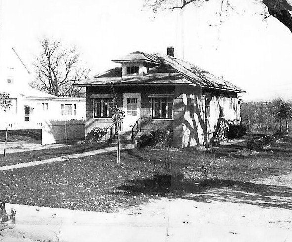 Black-and-white photo of a small single-story bungalow with a front porch, steps, lawn, and leafless trees in the yard.
