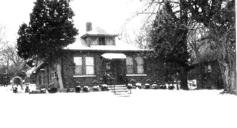 Snow-covered brick bungalow with front steps, shrubs and tall trees on either side.