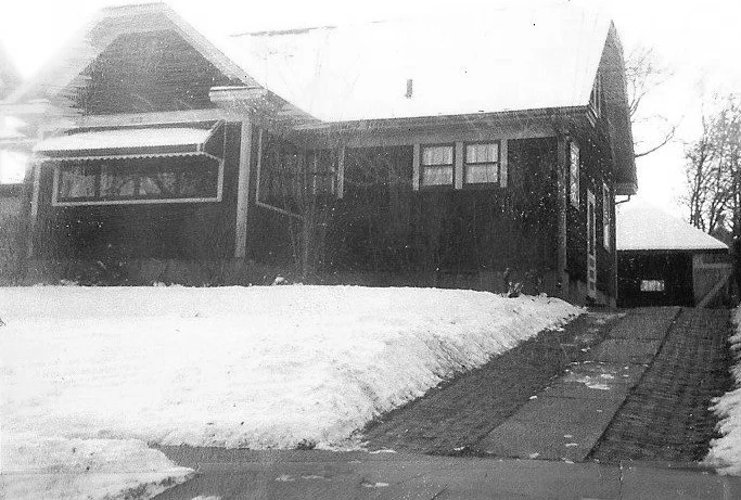 Black-and-white photo of a single-story bungalow with a snow-covered front yard and a driveway leading to a detached garage.
