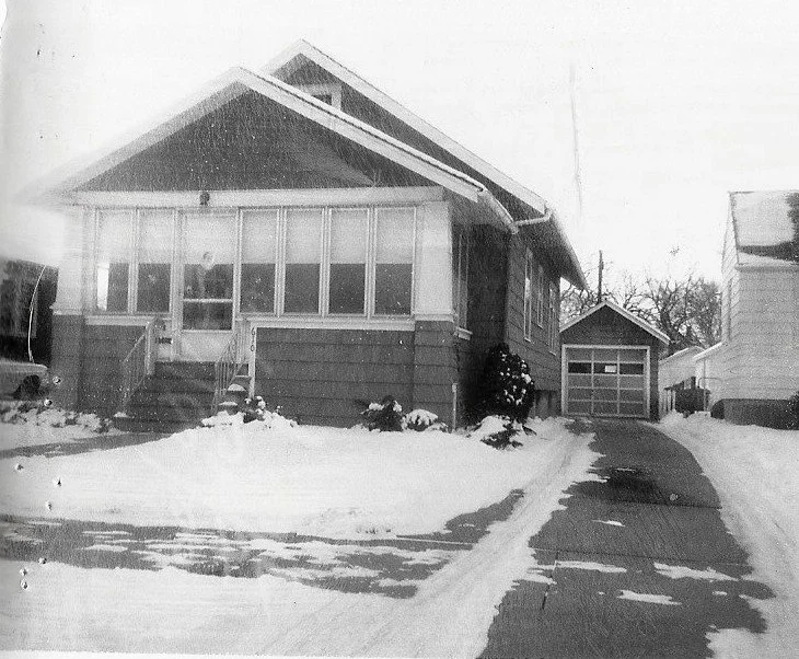 Black-and-white photo of a single-story bungalow with an enclosed front porch, snow-covered yard, and a driveway leading to a detached garage.