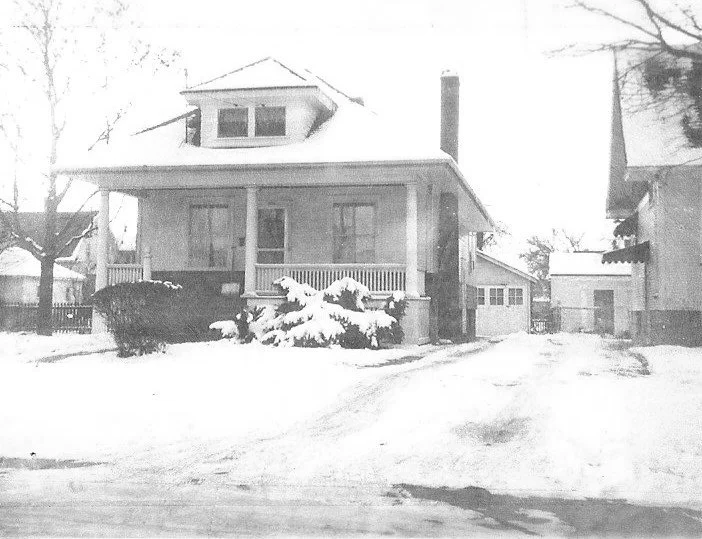 Snow-covered bungalow with a front porch and shrubs, driveway leading to a detached garage, flanked by neighboring houses.