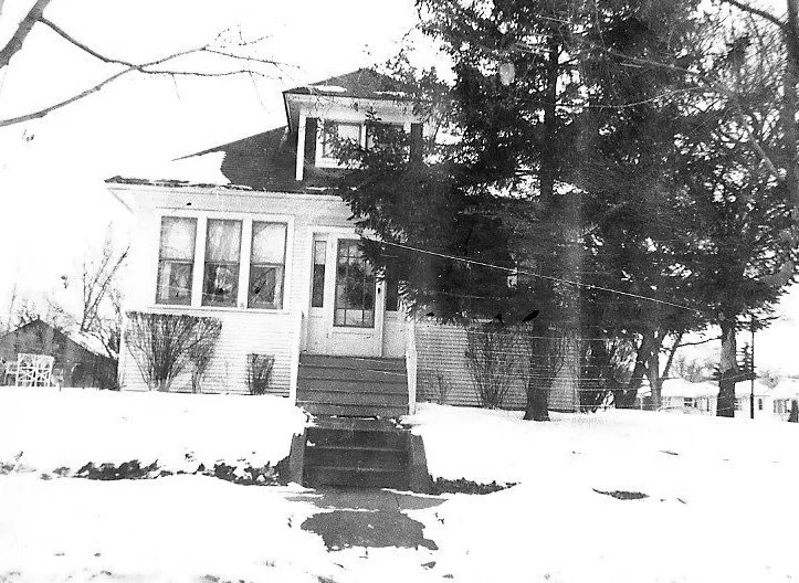 Black-and-white photo of a two-story house with front steps and a large evergreen tree, set in a snowy yard.