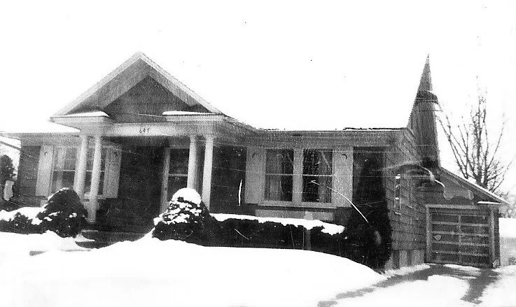 Black-and-white photo of a single-story bungalow with a columned front porch, attached garage, and snow-covered yard and shrubs.